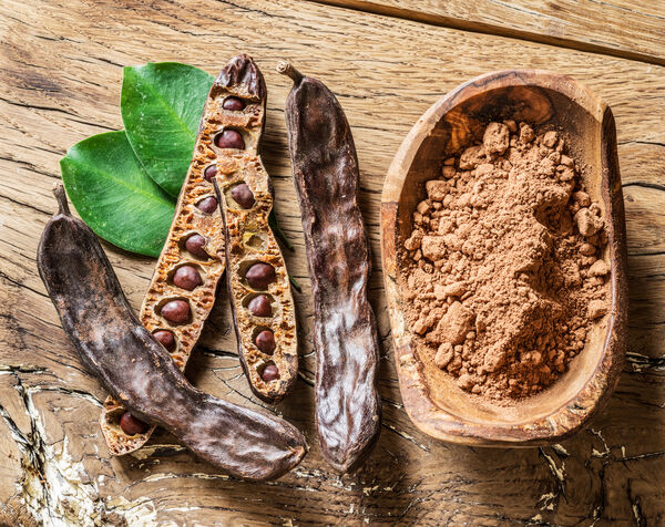 Carob-Pulver in Schale auf Holztisch neben Johannisbrotschoten
